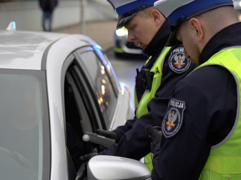 Dwaj policjanci w mundurach kontrolują dokumenty kierowcy w samochodzie osobowym na ruchliwej ulicy.