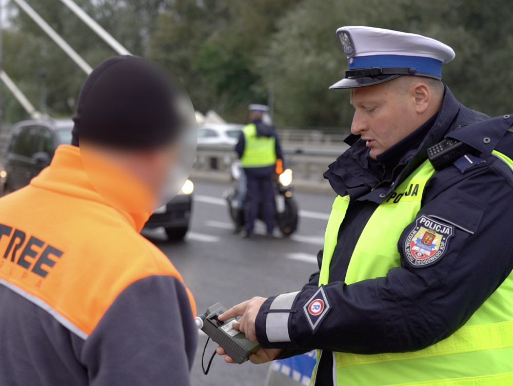 Policjant w kamizelce odblaskowej kontroluje mężczyznę w pomarańczowej odzieży roboczej na drodze.