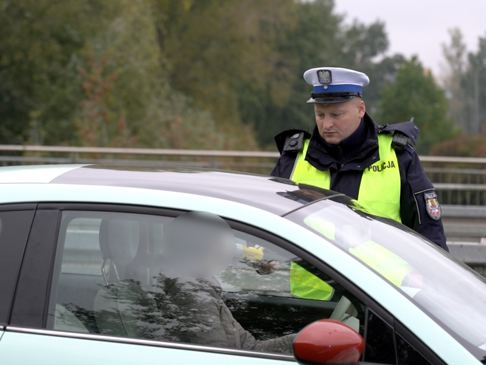 Policjant w kamizelce odblaskowej rozmawia z kierowcą w samochodzie na drodze.