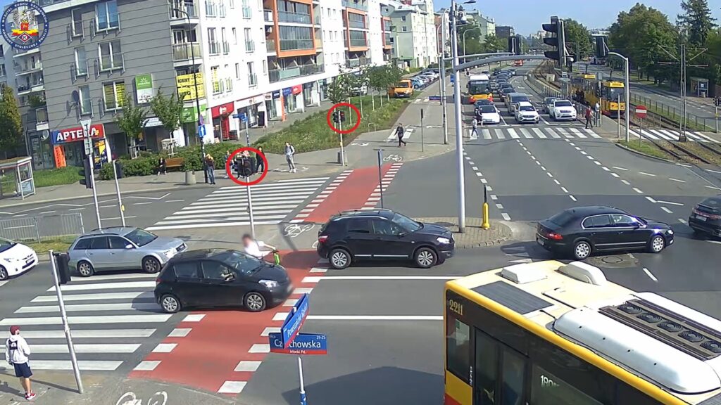 Grupa osób stojąca na chodniku przy przejściu dla pieszych, obok ruchliwej ulicy, w tle samochody i tramwaj.