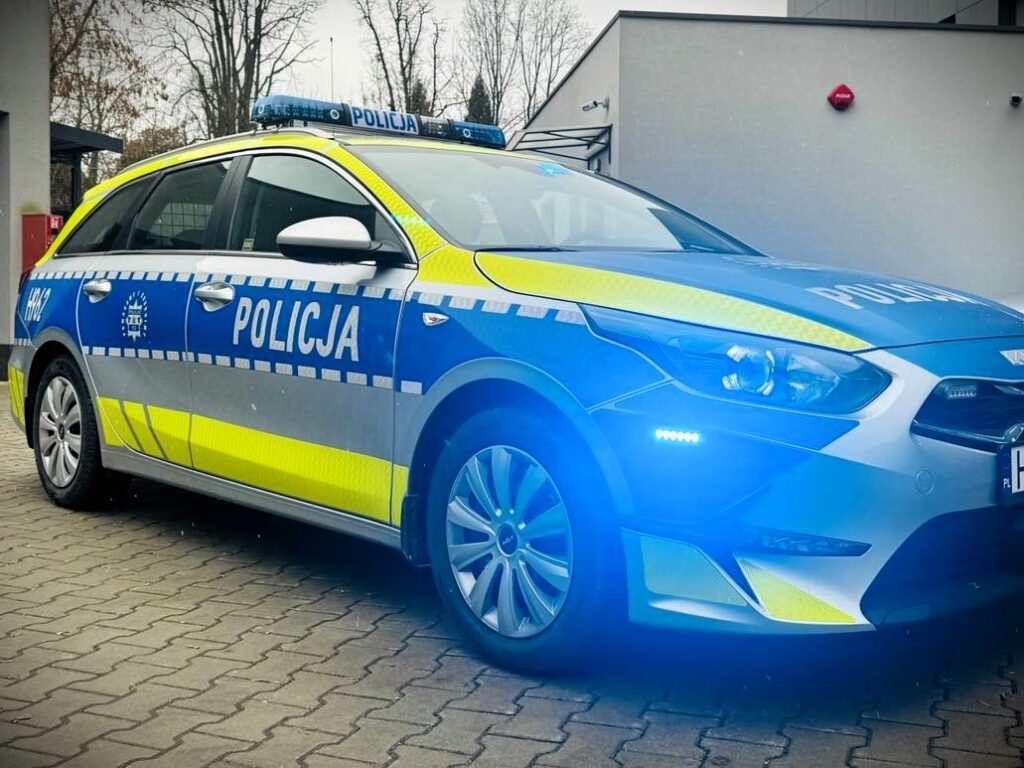 Radiowóz policyjny z niebieskimi i żółtymi oznaczeniami zaparkowany na kostce brukowej przed budynkiem.