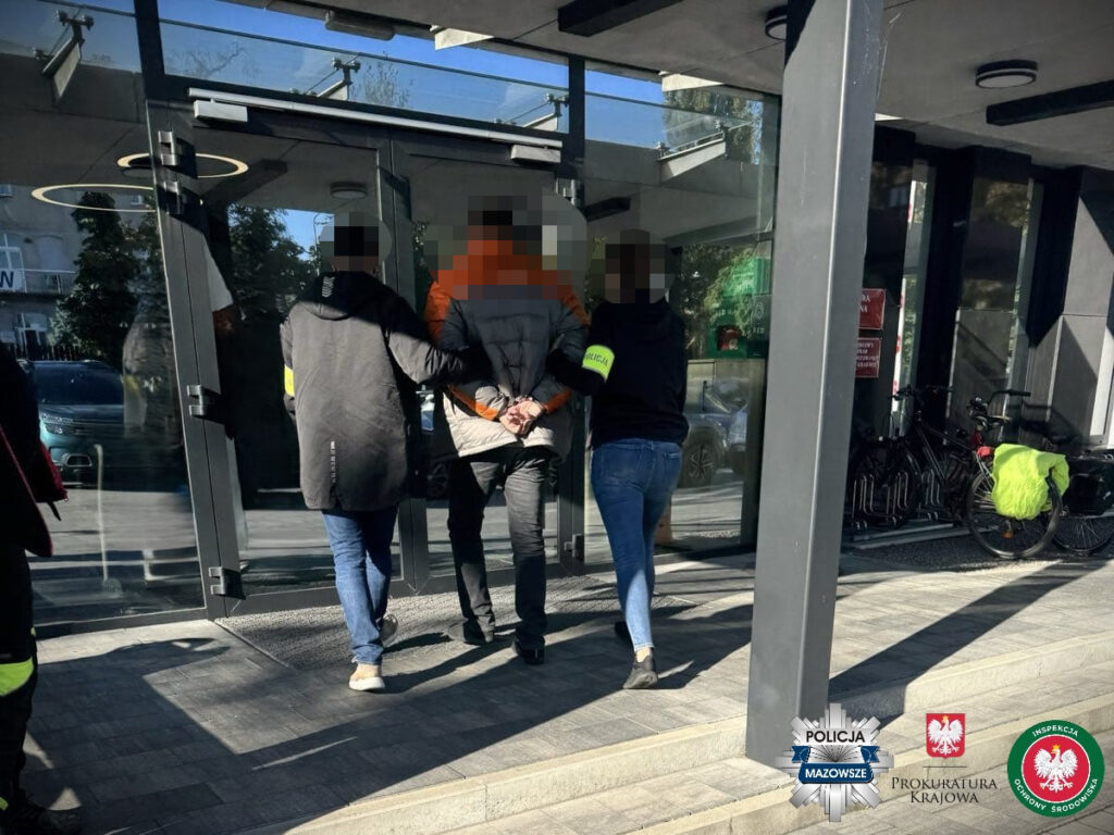 Mężczyzna w szarej kurtce prowadzony przez funkcjonariuszy policji do budynku z przeszkloną fasadą.