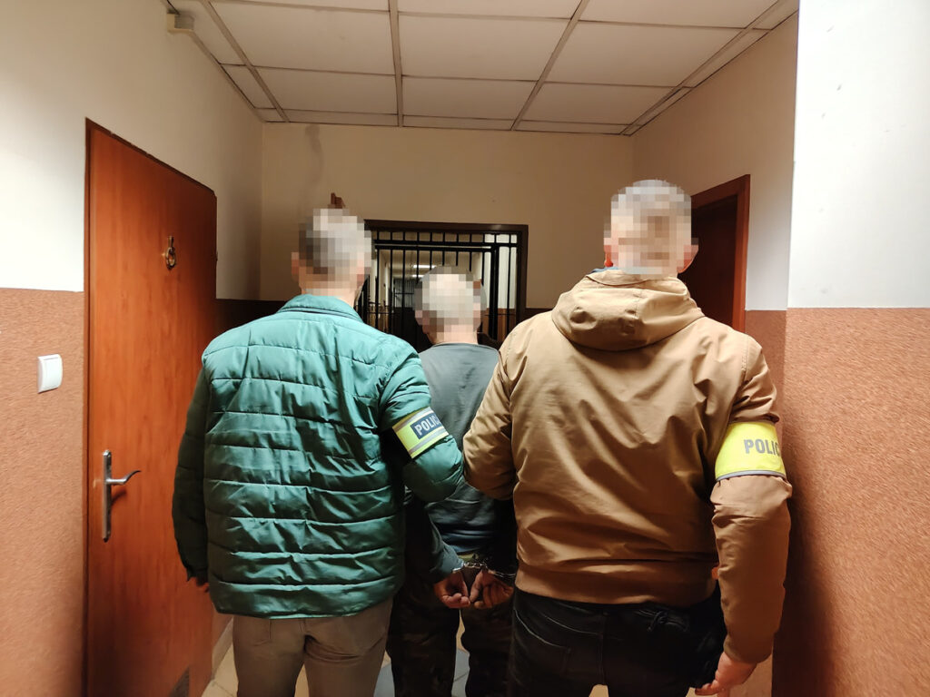 Trzech funkcjonariuszy policji prowadzi zatrzymanego mężczyznę korytarzem w budynku policyjnym.