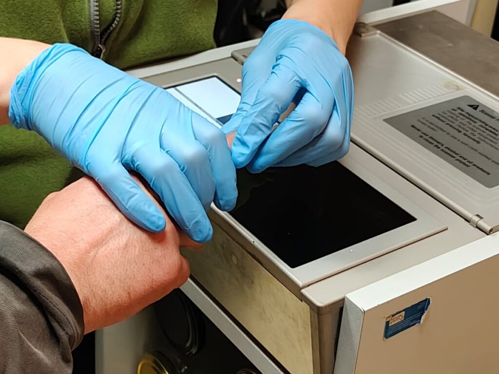 Osoba w niebieskich rękawiczkach dotyka ręki innej osoby, trzymając ją nad urządzeniem z ekranem dotykowym.