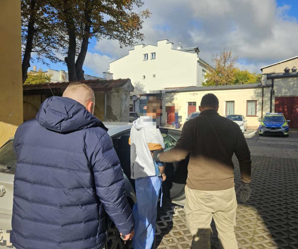 Mężczyzna w białej odzieży medycznej jest eskortowany przez dwóch mężczyzn w kierunku samochodu na parkingu.