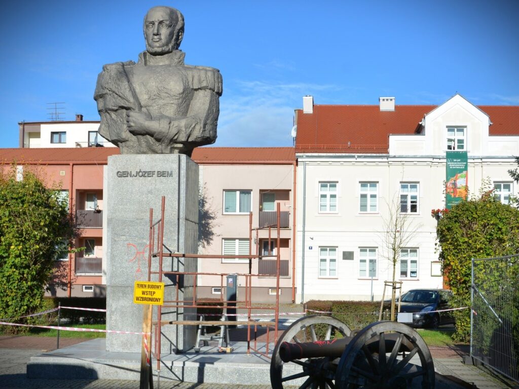 Pomnik gen. Józefa Bema w otoczeniu budynków mieszkalnych, z widocznymi zabezpieczeniami budowy i armatą w tle.