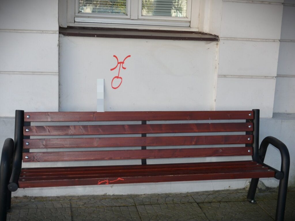 Czerwony graffiti w kształcie znaku na białej ścianie obok drewnianej ławki w miejskim otoczeniu.