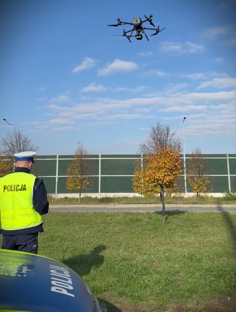 Policjant w kamizelce odblaskowej kontroluje drona unoszącego się nad terenem z zieloną trawą i drzewami.