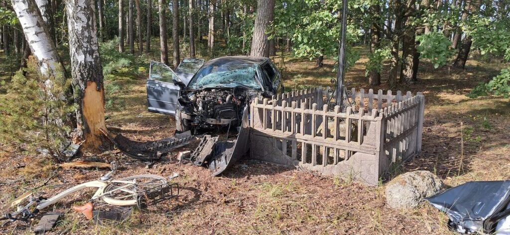 Uszkodzony samochód osobowy stoi w lesie obok drewnianego ogrodzenia, z otwartymi drzwiami i uszkodzoną maską.