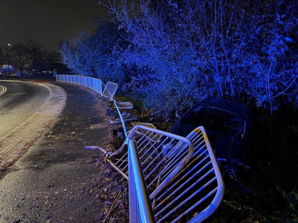 Złomowany samochód leży w rowie obok zakrętu, otoczony uszkodzonymi barierkami, w nocnym otoczeniu z niebieskim oświetleniem.