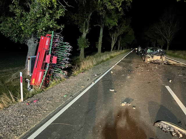 Zniszczony samochód leży na boku na drodze, obok przewrócony sprzęt rolniczy, otoczenie nocne z drzewami w tle.
