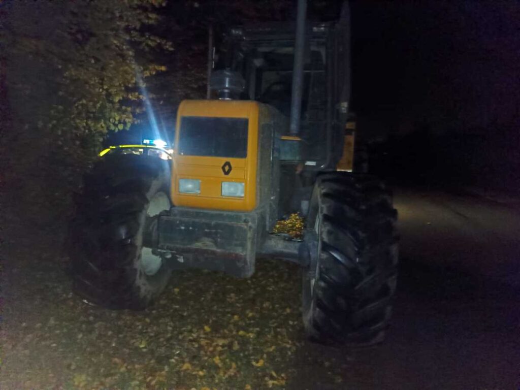 Traktor z pomarańczową kabiną stoi na drodze w nocy, w tle widoczna niebieska sygnalizacja.