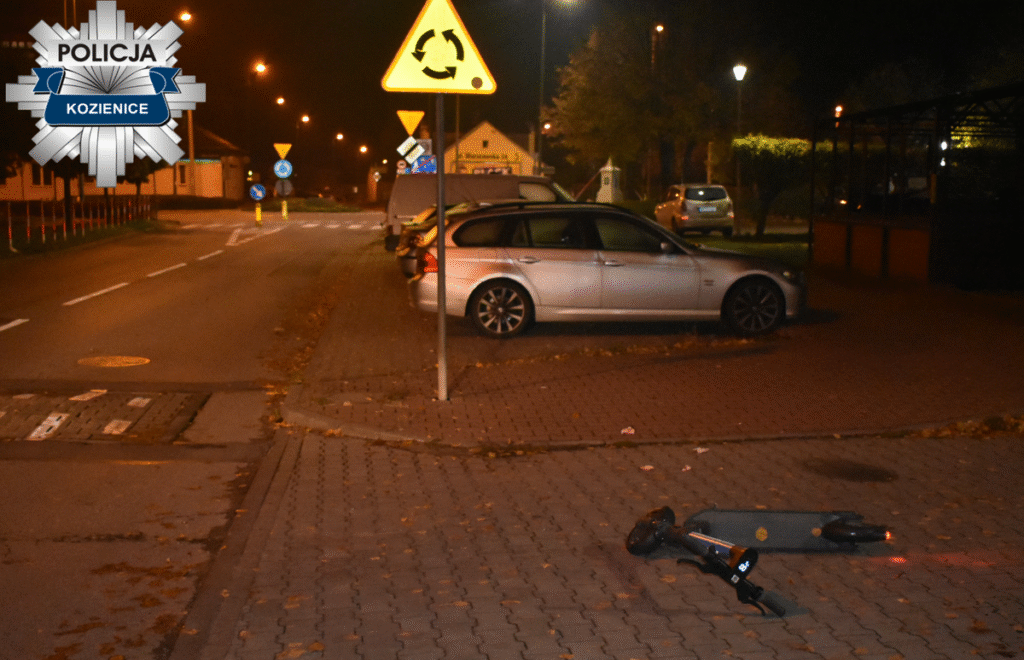 Na ulicy w nocy leży przewrócona hulajnoga elektryczna obok zaparkowanych samochodów. W tle widoczne znaki drogowe.