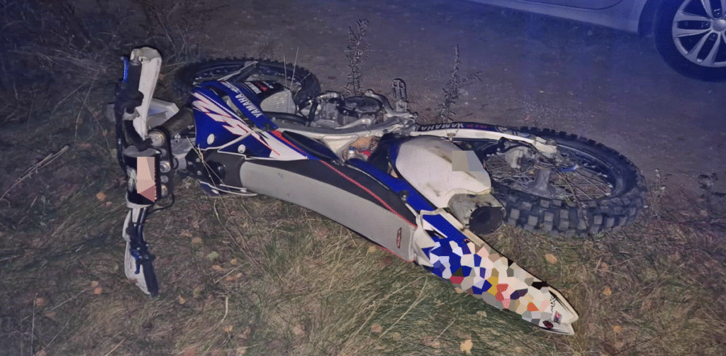 Motocykl terenowy leży na boku na trawie, obok widoczna jest droga i samochód w tle.