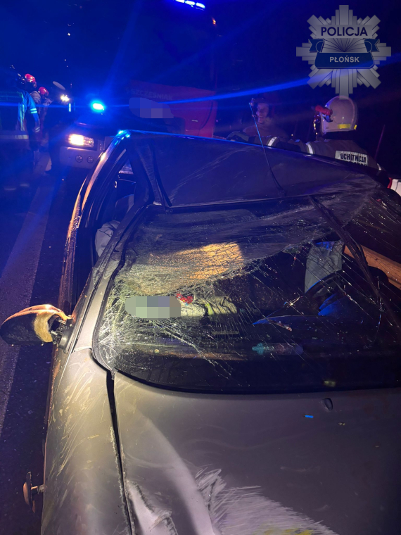 Uszkodzony samochód z rozbitą szybą dachową, otoczony przez strażaków i policję w nocy.