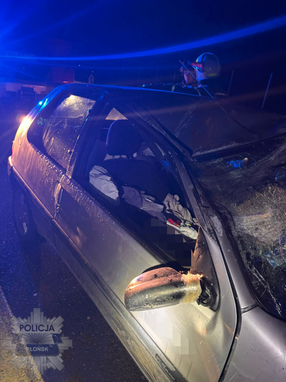 Uszkodzony samochód osobowy z wybitym oknem na drodze, obok stojący funkcjonariusz policji z latarką.