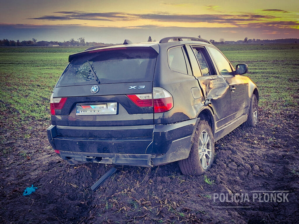 Czarny samochód osobowy marki BMW X3 utknął w polu, z widocznymi uszkodzeniami na boku. W tle widać zielone uprawy i zachodzące słońce.