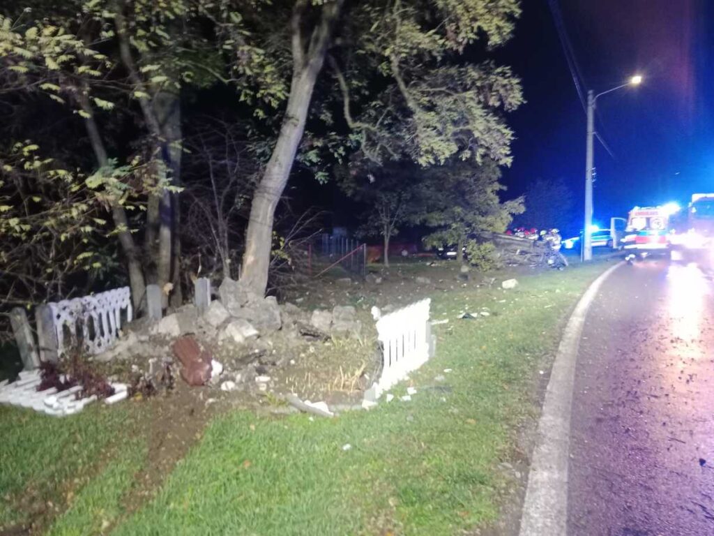 Zniszczony płot i fragmenty gruntu przy drodze, obok wozów strażackich i ambulansu w ciemnej scenerii nocnej.