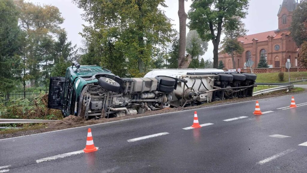 Przewrócony pojazd ciężarowy leży na poboczu drogi, otoczony pachołkami drogowymi, w tle widoczny kościół.