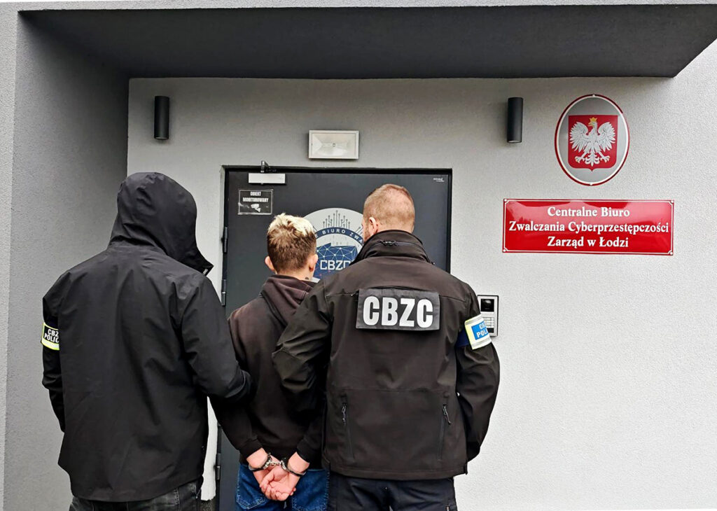 Mężczyzna w kajdankach prowadzony przez dwóch funkcjonariuszy przed budynkiem Centralnego Biura Zwalczania Cyberprzestępczości.