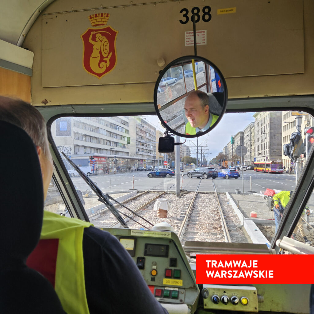 Kierowca tramwaju w kamizelce odblaskowej patrzy w lusterku, prowadząc pojazd na torach w mieście.
