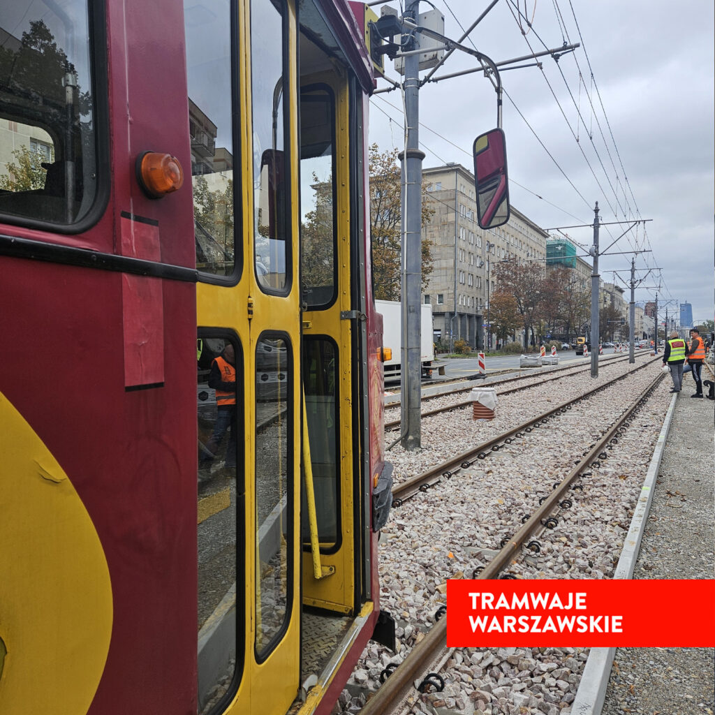 Tramwaj w kolorze bordowym i żółtym stoi na torach, obok pracownicy w kamizelkach odblaskowych prowadzą prace na ulicy.