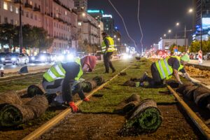 Pracownicy w kamizelkach odblaskowych układają trawniki wzdłuż torów tramwajowych w nocy.