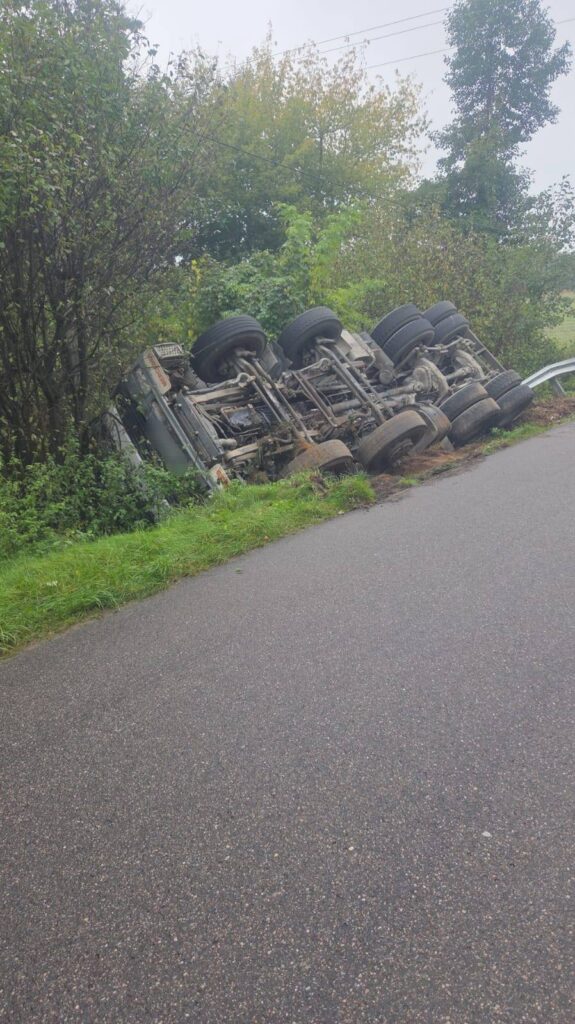 Przewrócony ciężarówka leżąca na poboczu drogi, otoczona zielenią i drzewami.