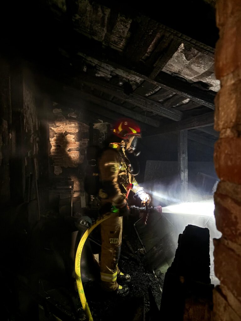 Strażak w hełmie i specjalnym ubraniu gasi pożar w zniszczonym pomieszczeniu, oświetlając je latarką.