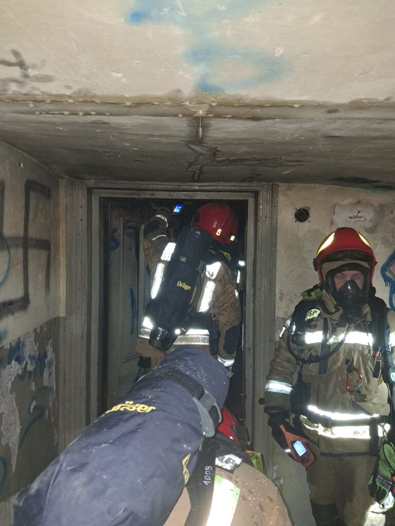 Grupa strażaków w pełnym wyposażeniu przeszukuje zniszczony korytarz w opuszczonym budynku.