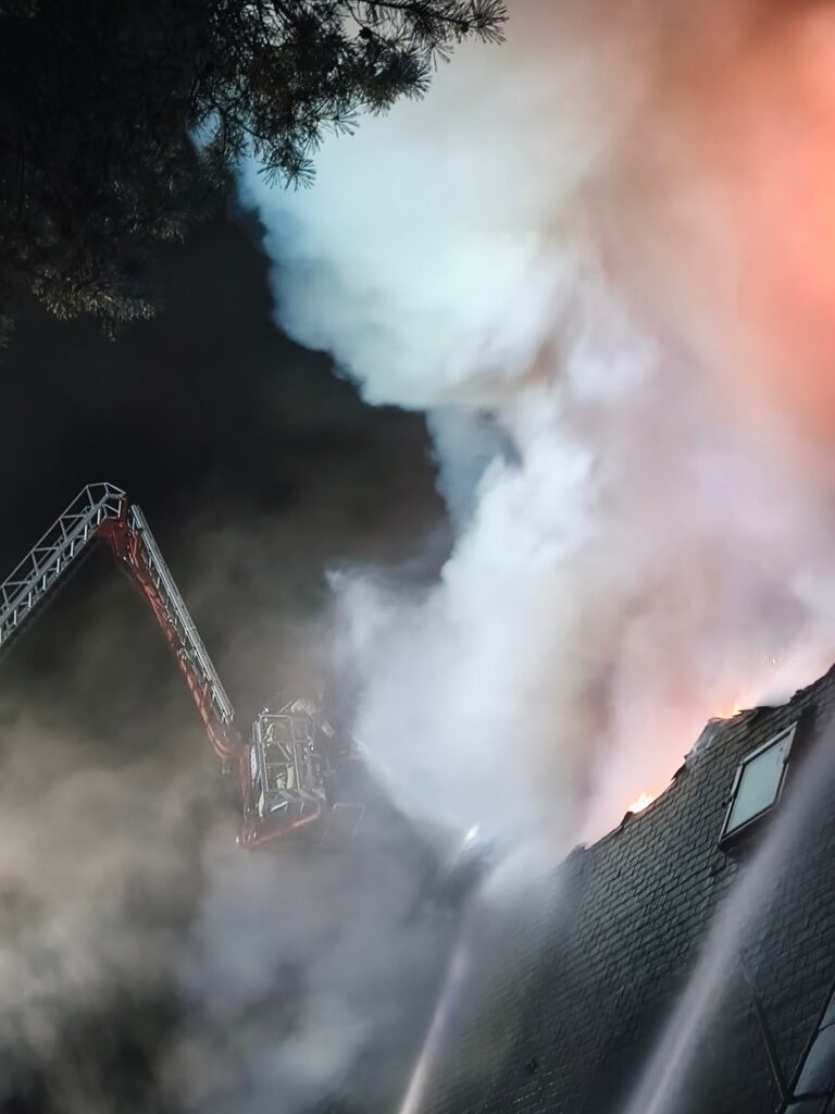 Strażak na podnośniku gaśniczym polewa wodą płonący dach budynku, otoczony gęstym dymem.
