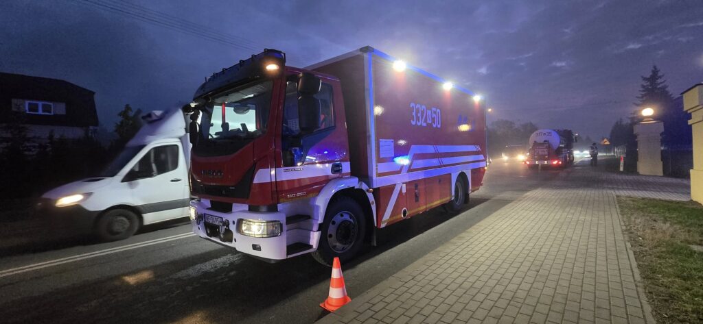 Czerwony wóz strażacki z włączonymi światłami sygnalizacyjnymi stoi na drodze, obok przejeżdżają białe dostawcze auto.