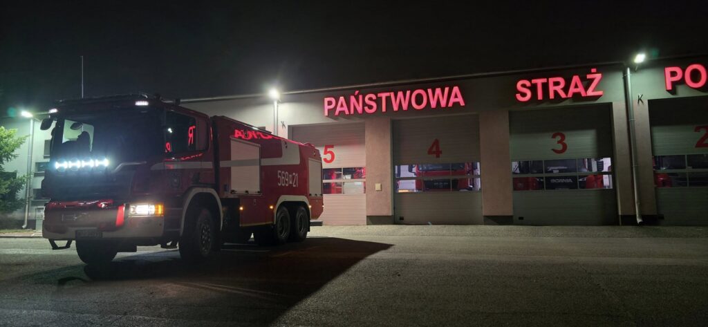 Czerwony wóz strażacki zaparkowany przed remizą strażacką oświetloną neonowymi napisami.