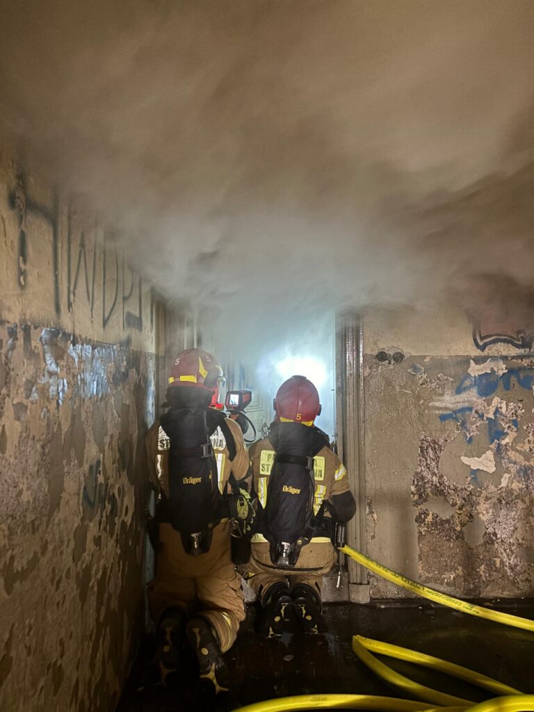 Dwaj strażacy w hełmach i sprzęcie ochronnym, klęczący w zadymionym korytarzu, monitorują sytuację z kamerą.