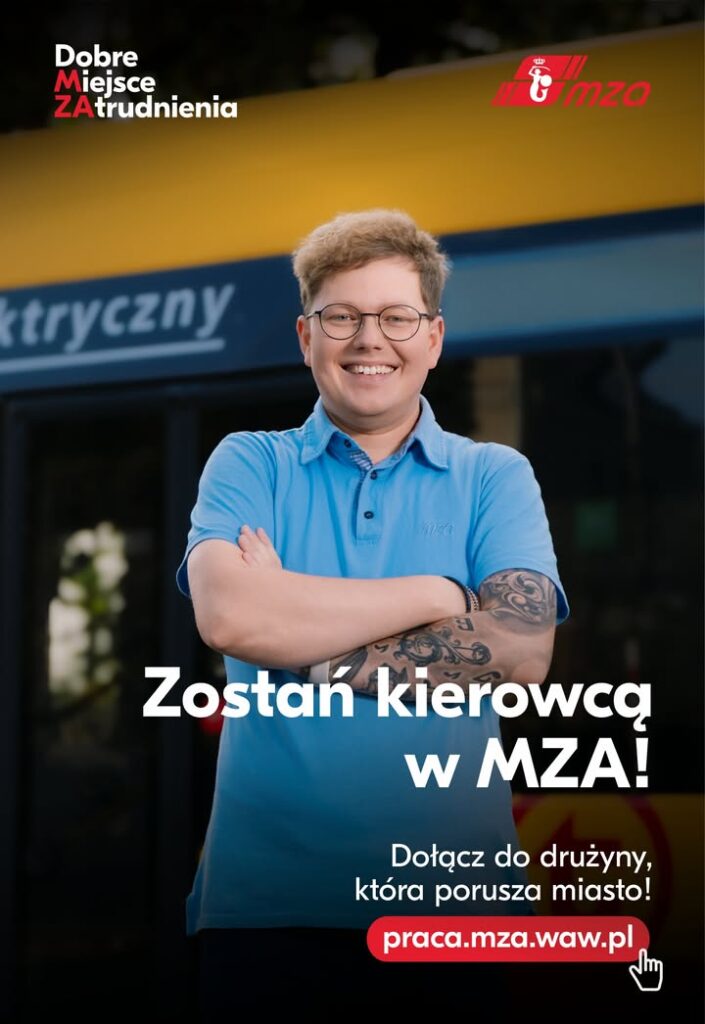 Młody mężczyzna z tatuażami, uśmiechając się, stoi z założonymi rękami przed autobusem w tle.
