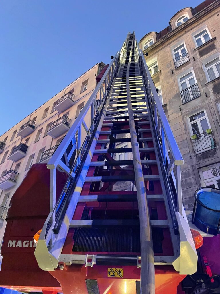 Wysoka drabina strażacka skierowana w stronę budynku z balkonami w wieczornym świetle.