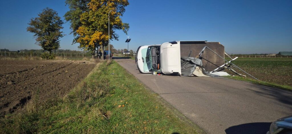 Przewrócony samochód dostawczy leży na drodze, obok pola uprawnego, w otoczeniu drzew i niebieskiego nieba.