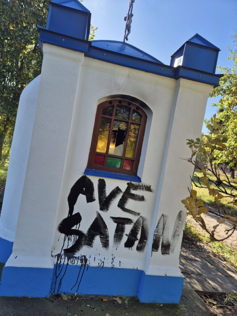 Kapliczka z kolorowymi witrażami, na której ścianie widnieje graffiti z napisem "AVE SATAN".