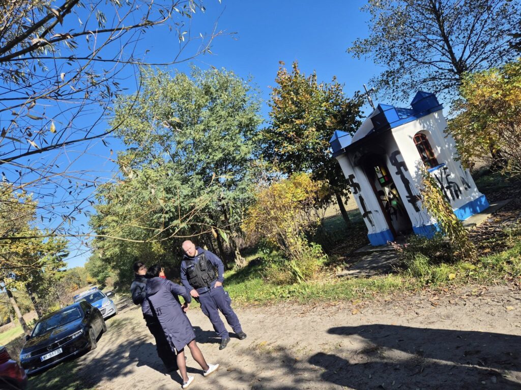 Dwóch policjantów rozmawia z kobietą na leśnej drodze obok małego, kolorowego budynku. W tle widać samochody i drzewa.