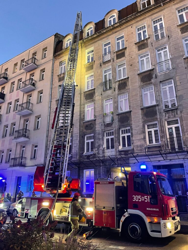 Strażacy przy użyciu wozu strażackiego z drabiną interweniują przy budynku mieszkalnym w godzinach wieczornych.
