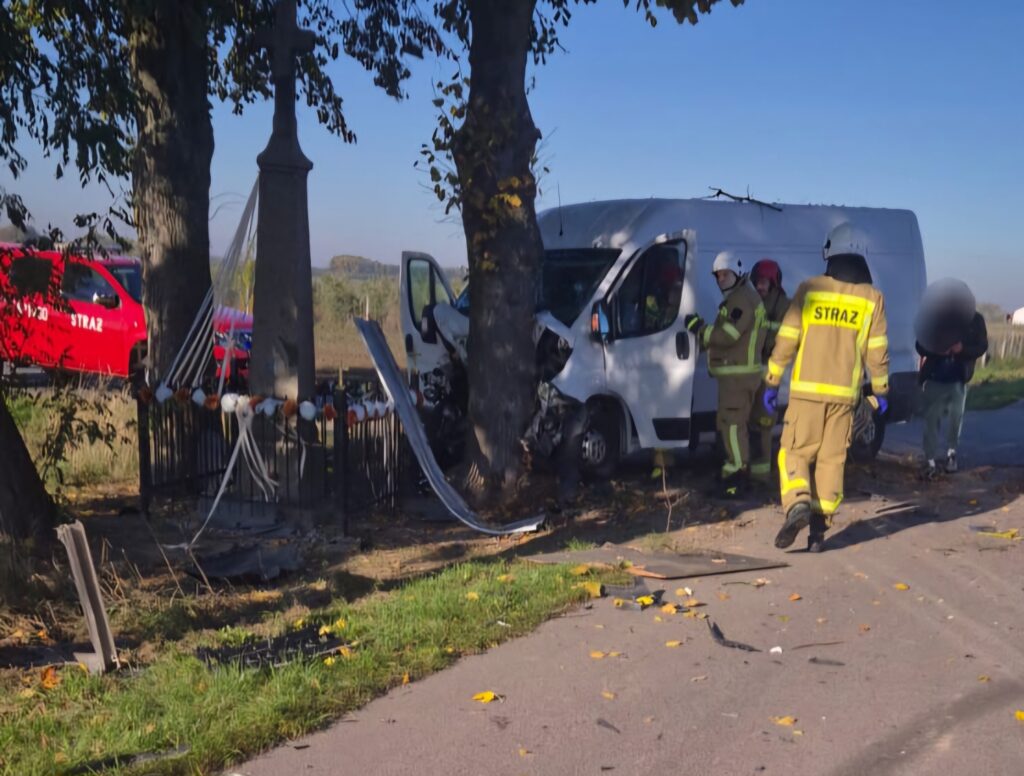Strażacy w odzieży ochronnej zabezpieczają miejsce wypadku przy drodze, obok uszkodzonego furgonu i pomnika.