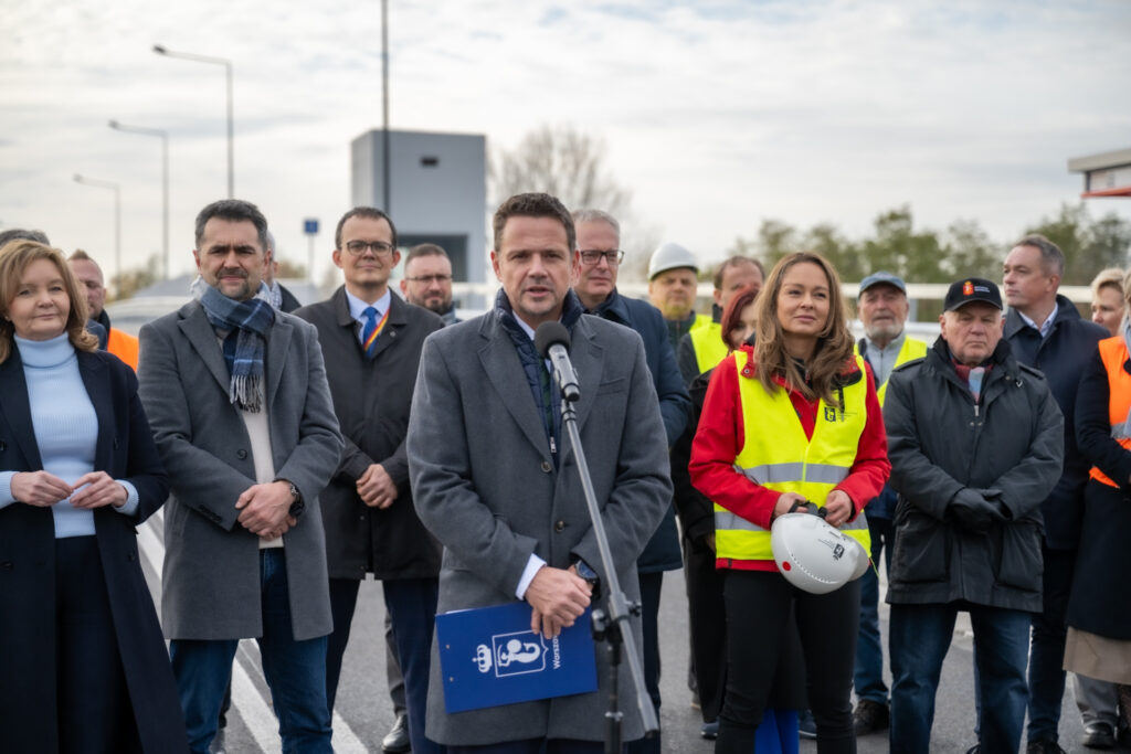 Mężczyzna w szarym płaszczu przemawia do zgromadzonego tłumu w kamizelkach odblaskowych na otwartej przestrzeni.