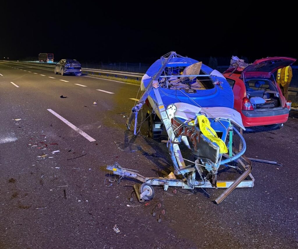 Zniszczony niebieski pojazd leży na jezdni obok czerwonego samochodu, otoczone przez rozbite elementy i szczątki.