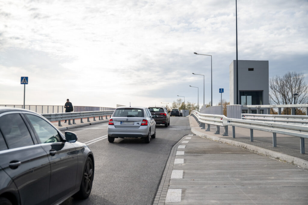 Samochody przejeżdżają przez nowoczesny most, obok spacerujący mężczyzna z torbą w dłoni.