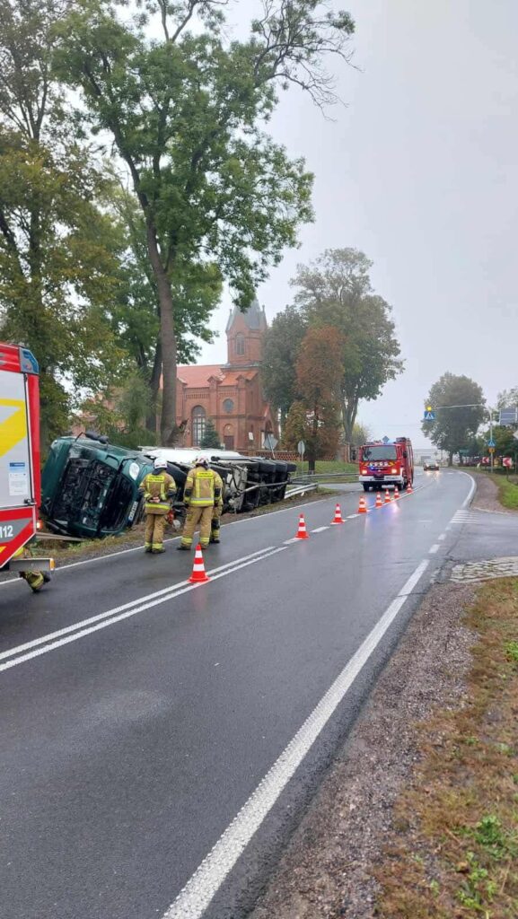 Strażacy pracują nad przewróconym pojazdem na drodze, w tle widoczny kościół w deszczowej aurze.