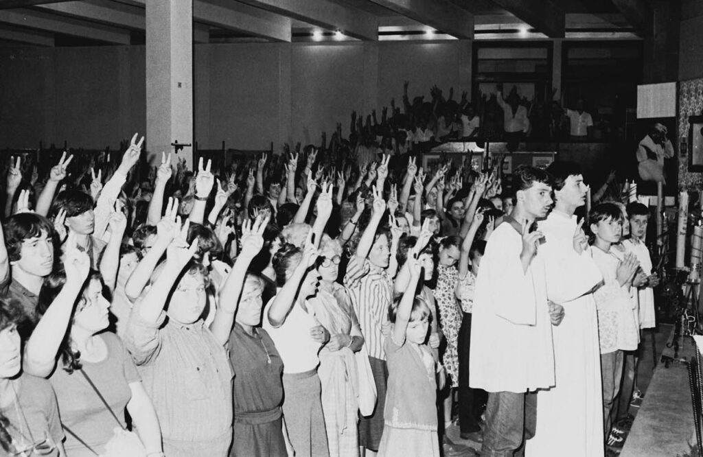 Młodzież w strojach codziennych podnosi ręce w geście zwycięstwa podczas wydarzenia religijnego w dużej sali.