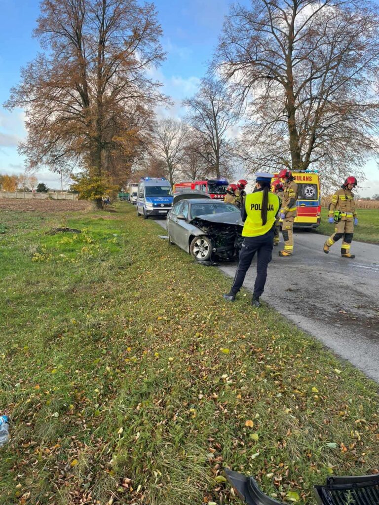 Kierowca w samochodzie osobowym z uszkodzoną przednią częścią, obok interweniujący strażacy i policja na drodze.