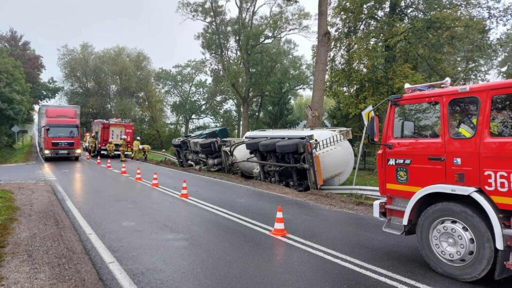 Czerwony wóz strażacki oraz ciężarówka z przewróconą naczepą na drodze, wokół rozstawione pachołki.