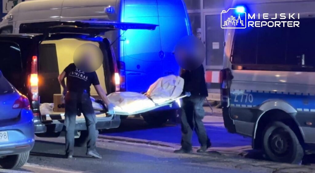 Mężczyźni w odzieży roboczej przenoszą zwłoki na noszach z furgonetki w oświetlonym nocą otoczeniu ulicznym.