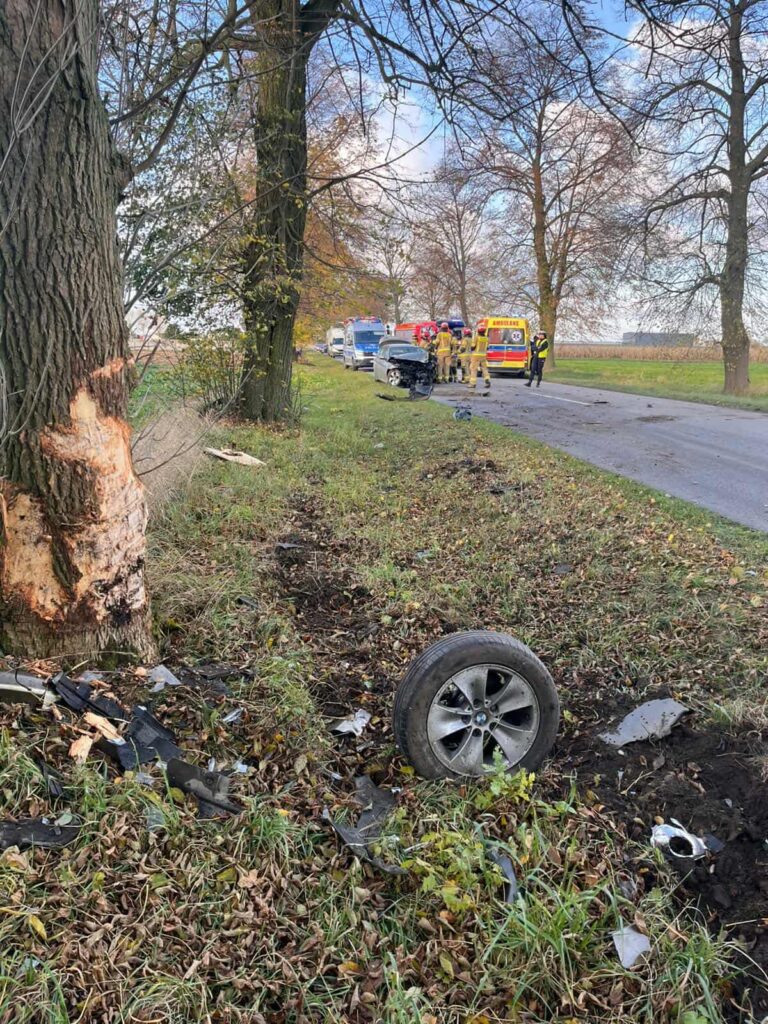 W miejscu wypadku samochodowego leży rozbity pojazd, obok którego pracują strażacy i ratownicy medyczni.